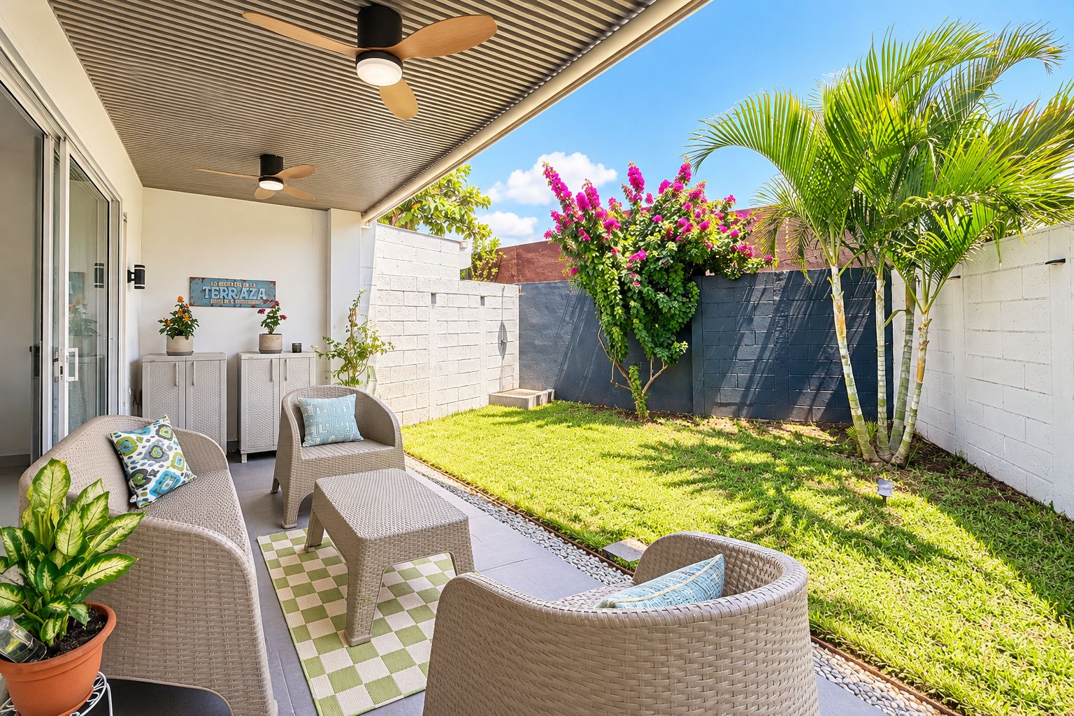 A covered patio area with wicker furniture, a checkered floor, and a small garden with a palm tree and flowering plants.