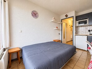 Ein kleines Zimmer mit einem Bett, einem kleinen Holzschemel, einer Wanduhr, einer Mikrowelle und einem Kühlschrank.