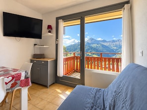 Ein Zimmer mit einer Couch, einem Tisch mit einer Patchworkdecke, einem an der Wand montierten Fernseher, einer kleinen Kommode und einem Balkon mit Bergblick.