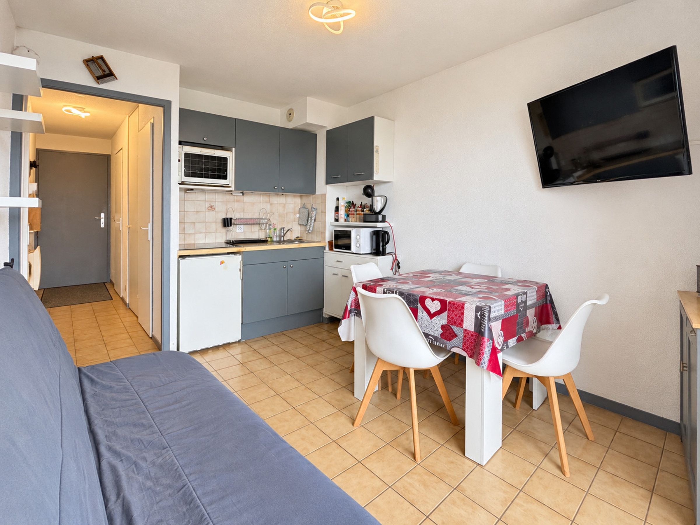 A compact kitchen with a dining area, a sofa, and a TV mounted on the wall.