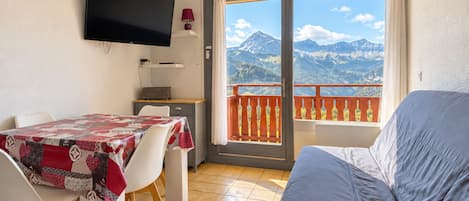 A room with a bed, a TV mounted on the wall, a table with chairs, and a balcony with a mountain view.