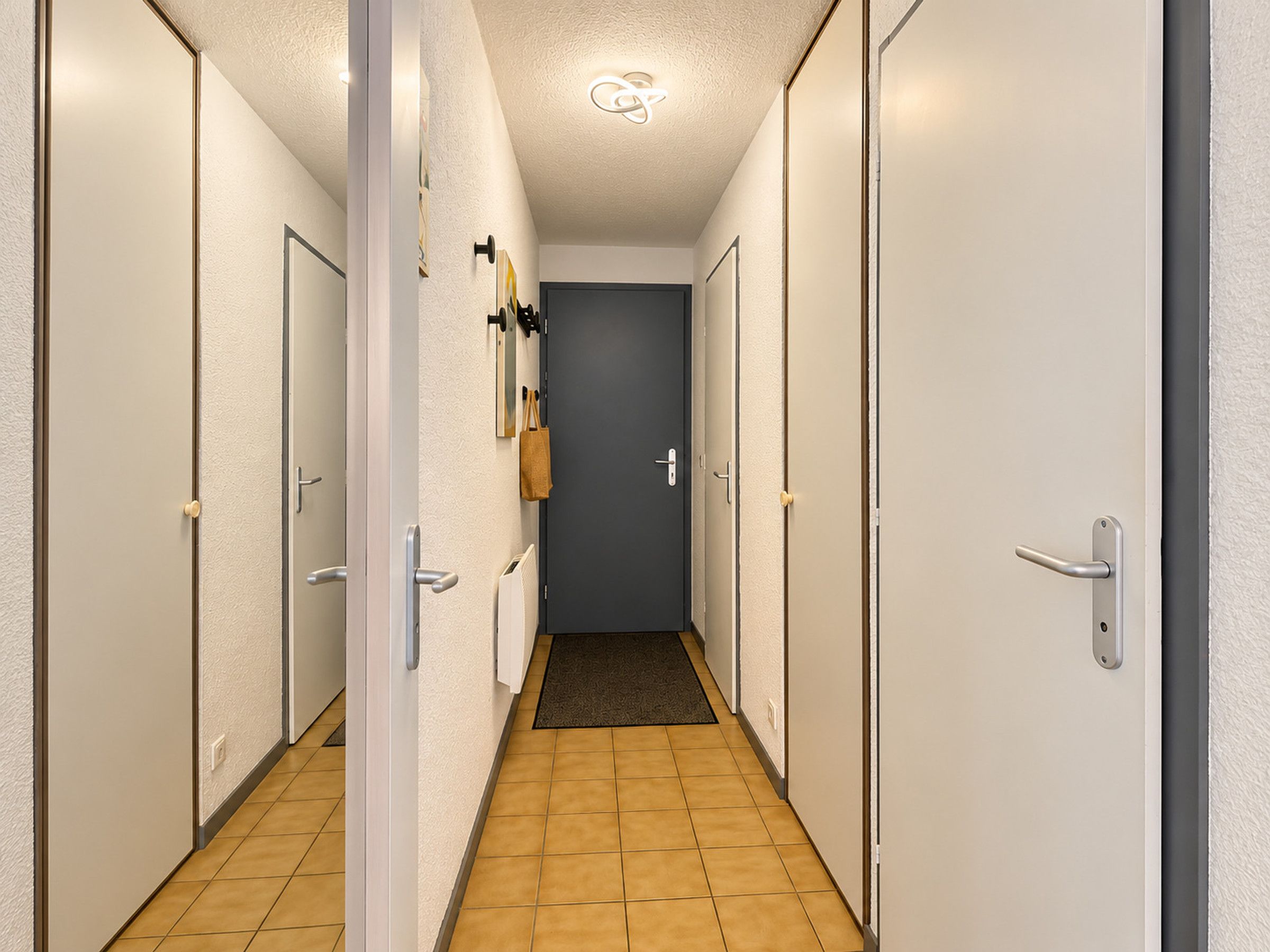 Un couloir avec des portes des deux côtés, une porte sombre au fond, et un mur de couleur claire avec un revêtement texturé.