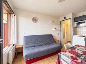 Quarto compacto com sofá cinza, uma pequena mesa de madeira, cozinha com micro-ondas e uma porta que dá para outro quarto.