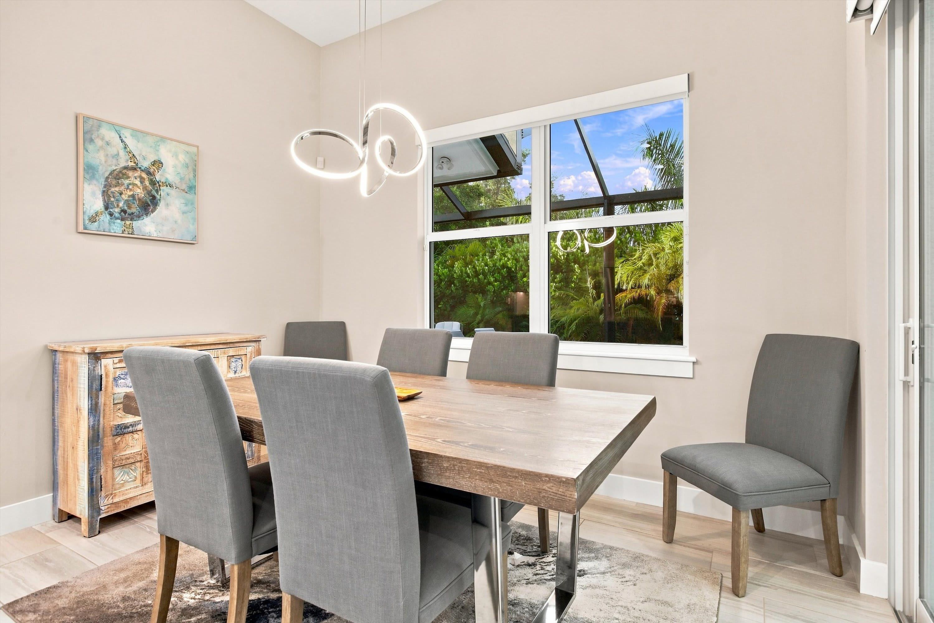 A dining area with a wooden table, grey upholstered chairs, a decorative cabinet, and a large window with a view of greenery.