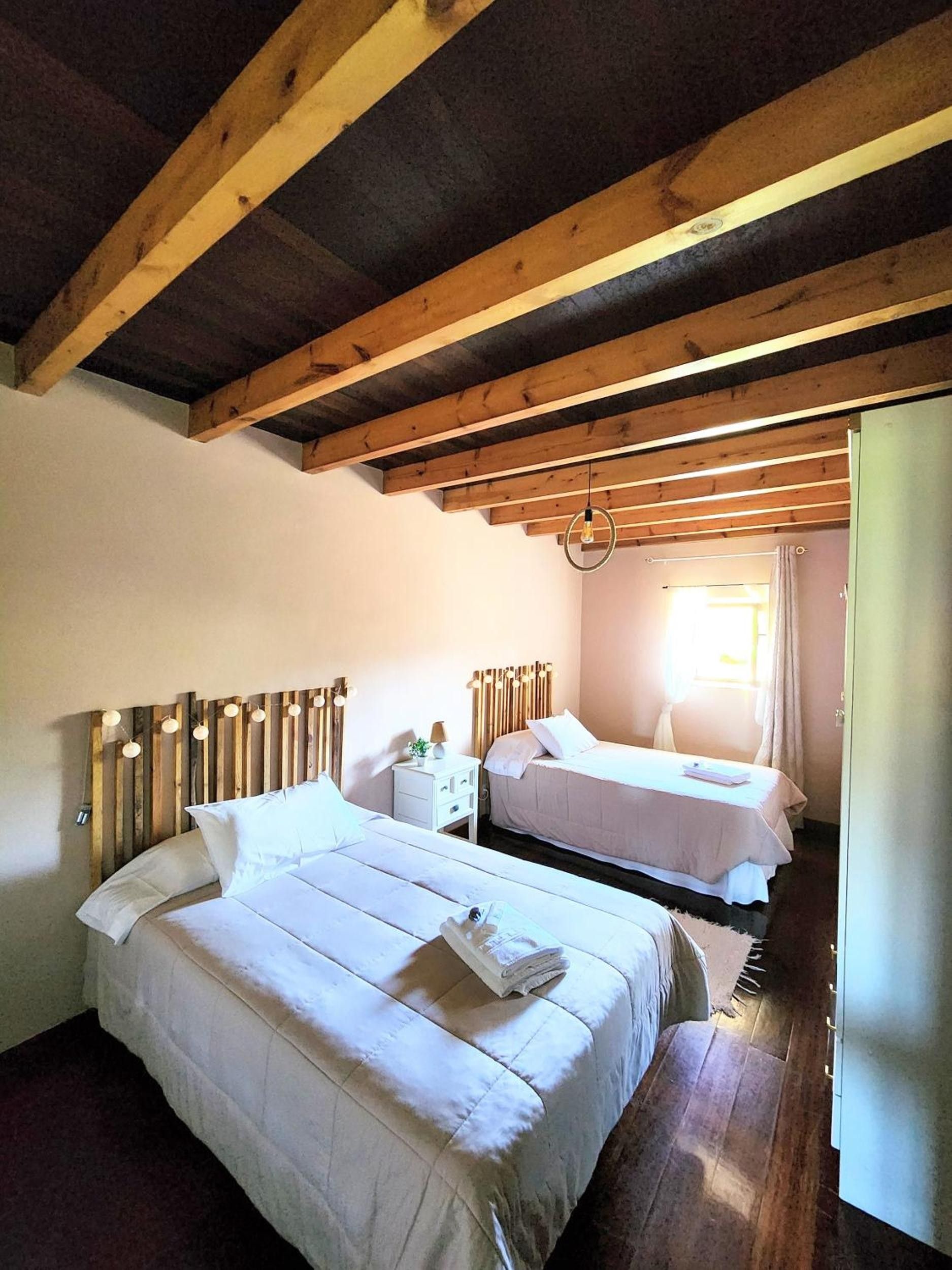 Une chambre avec deux lits, des poutres en bois au plafond, un luminaire suspendu et une fenêtre avec des rideaux.
