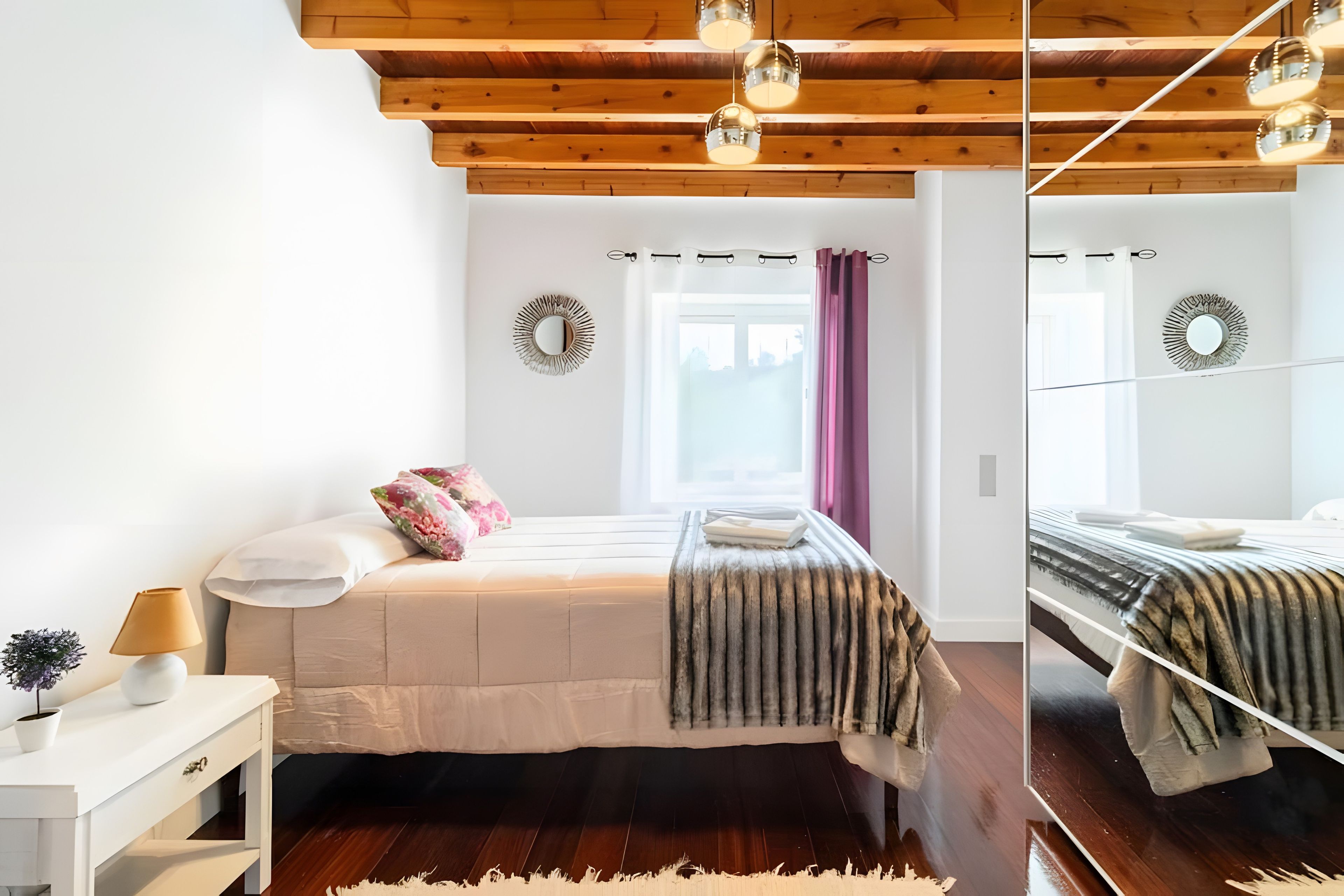 Een slaapkamer met een houten plafond, een bed met een witte dekbedovertrek, een nachtkastje met een lamp en een raam met gordijnen.