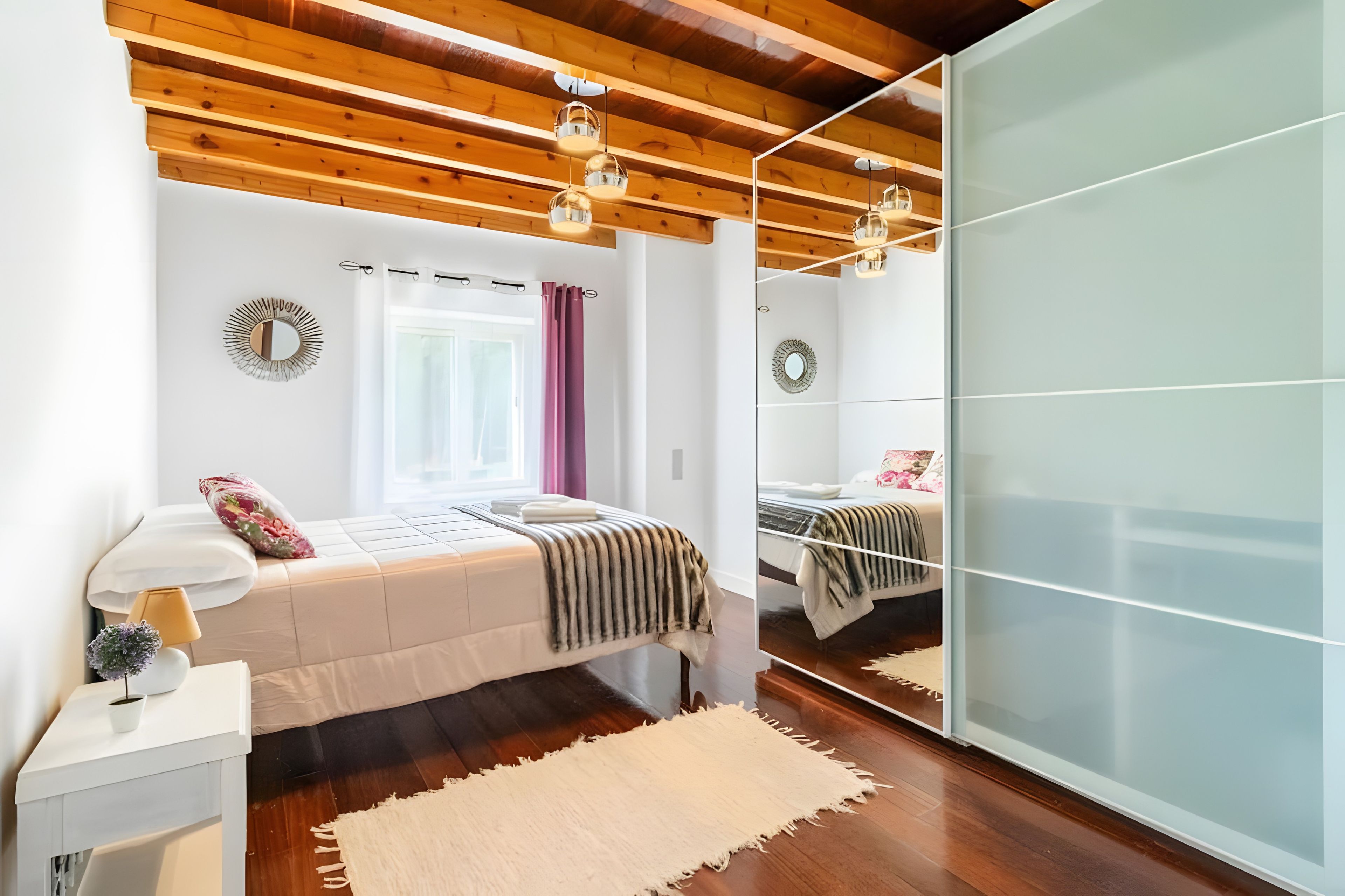 Een slaapkamer met een bed, nachtkastje, lamp, spiegel en een groot raam met gordijnen.