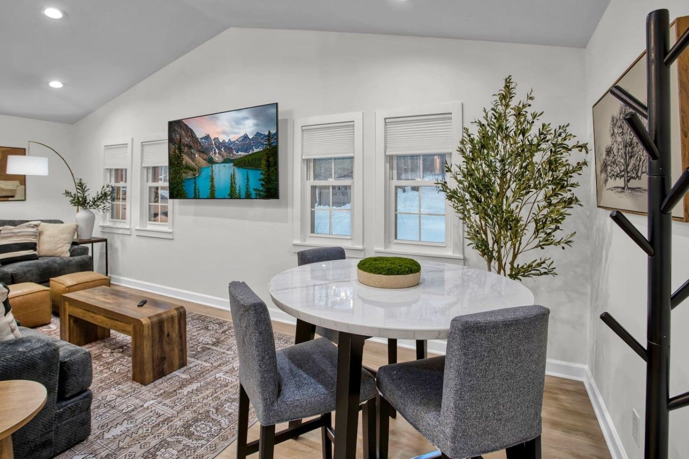 A modern living room with a round dining table, grey upholstered chairs, a sofa, and a large painting of a mountain landscape.