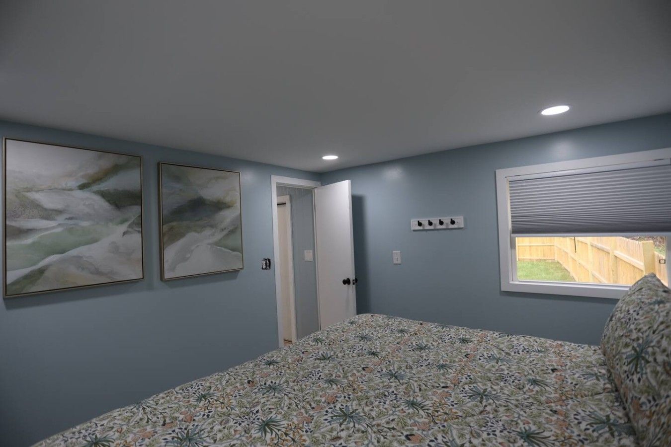 A bedroom with a bed, two wall-mounted paintings, a door, a window with blinds, and a power outlet.