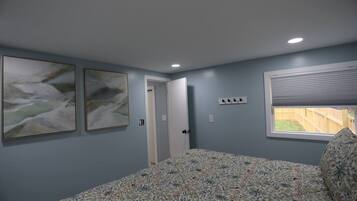 A bedroom with a bed, two wall-mounted paintings, a door, a window with blinds, and a power outlet.