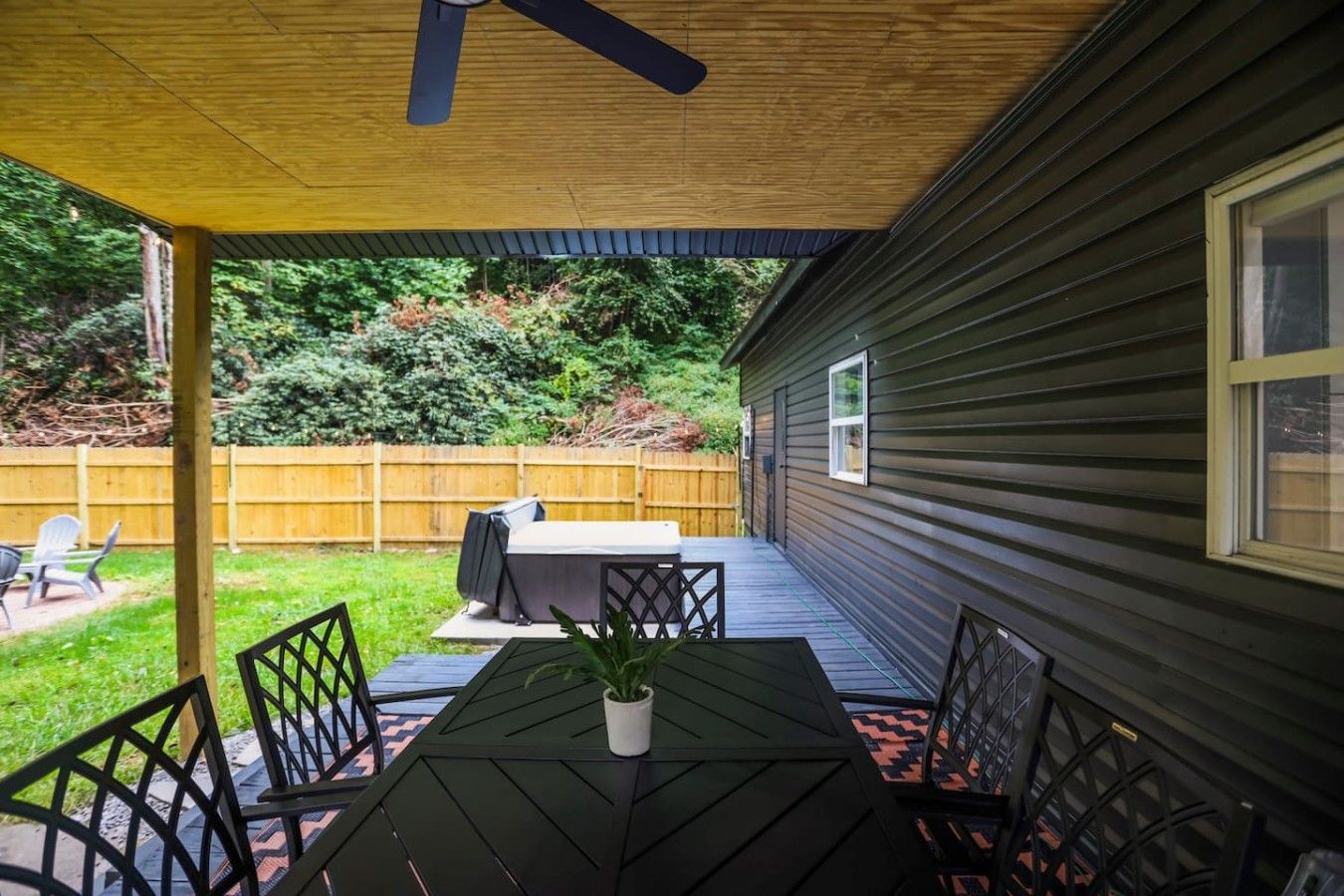 A covered patio with a table and chairs, a ceiling fan, and a view of a backyard with a wooden fence and greenery.