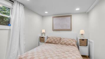 A bedroom with a bed, two bedside tables, a window with curtains, a wall-mounted artwork, and a ceiling light.