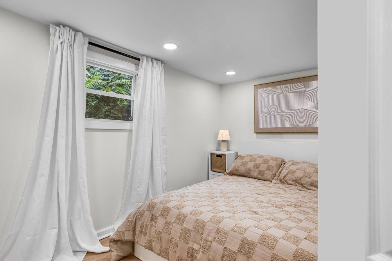 Ein Schlafzimmer mit einem Bett, einem Nachttisch, einer Lampe, einem Fenster mit Vorhängen und einem Wandbild.