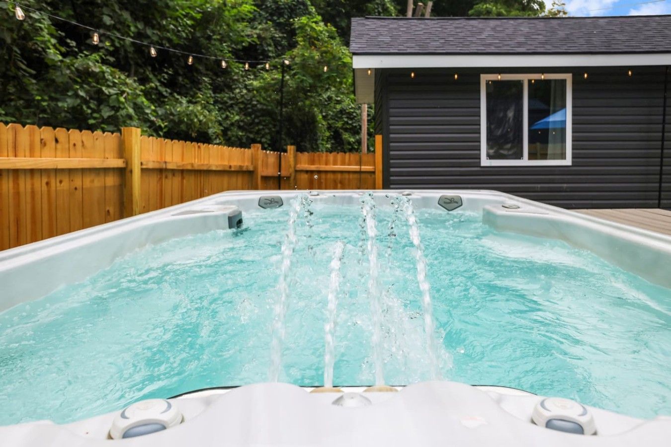 Een jacuzzi met waterstralen, omgeven door een houten schutting en een huis met een raam.