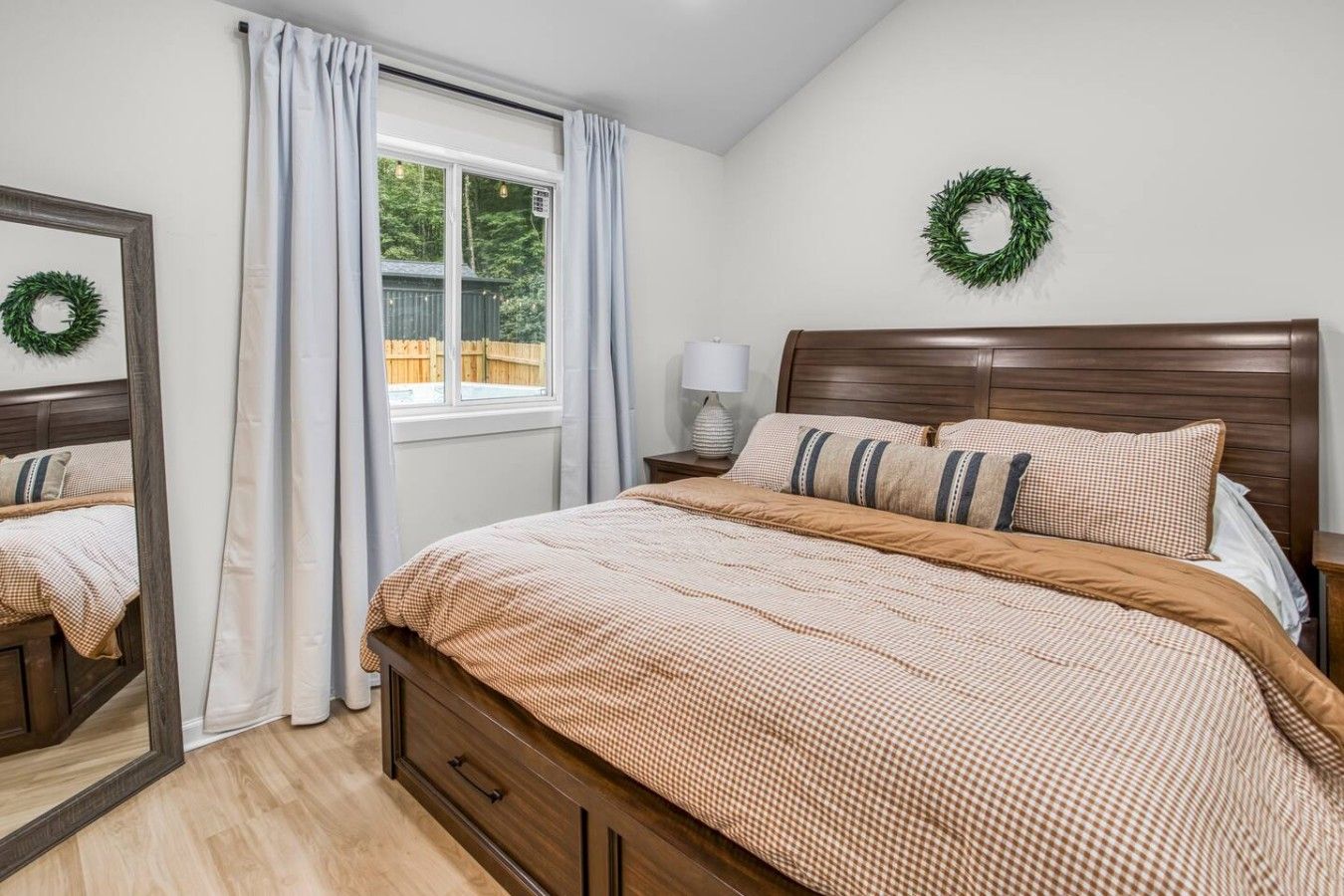 Un dormitorio con cama de madera, ventana con cortinas, mesita de noche con lámpara y una corona en la pared.