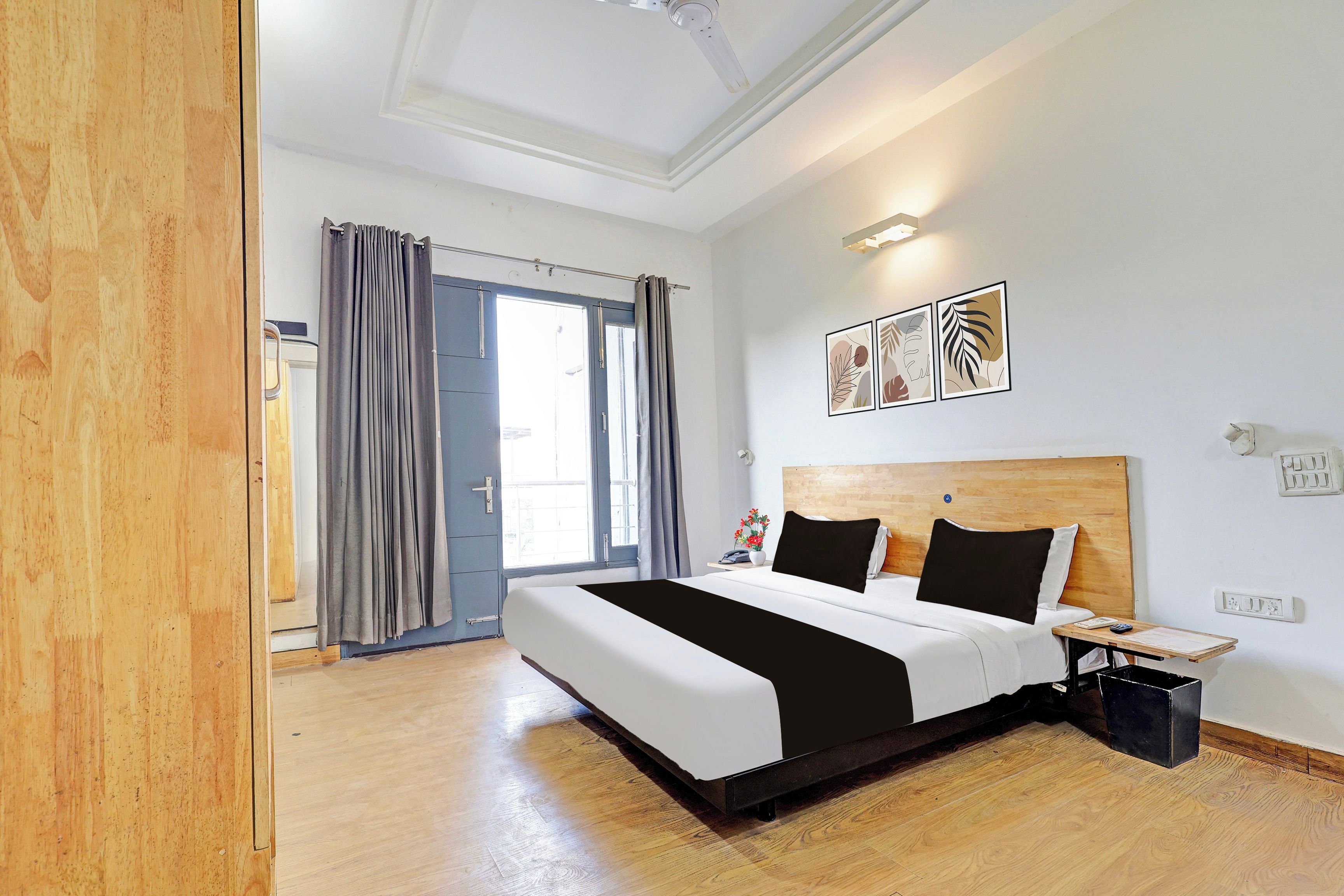 A hotel room with a large bed, a wooden headboard, a bedside table, a nightstand, a wall-mounted light, and a window with curtains.