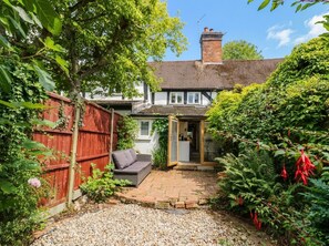 A residential property with a brick patio, a wooden fence, and a garden with various plants and a red flowering plant. - Lilac Cottage (Rugby)