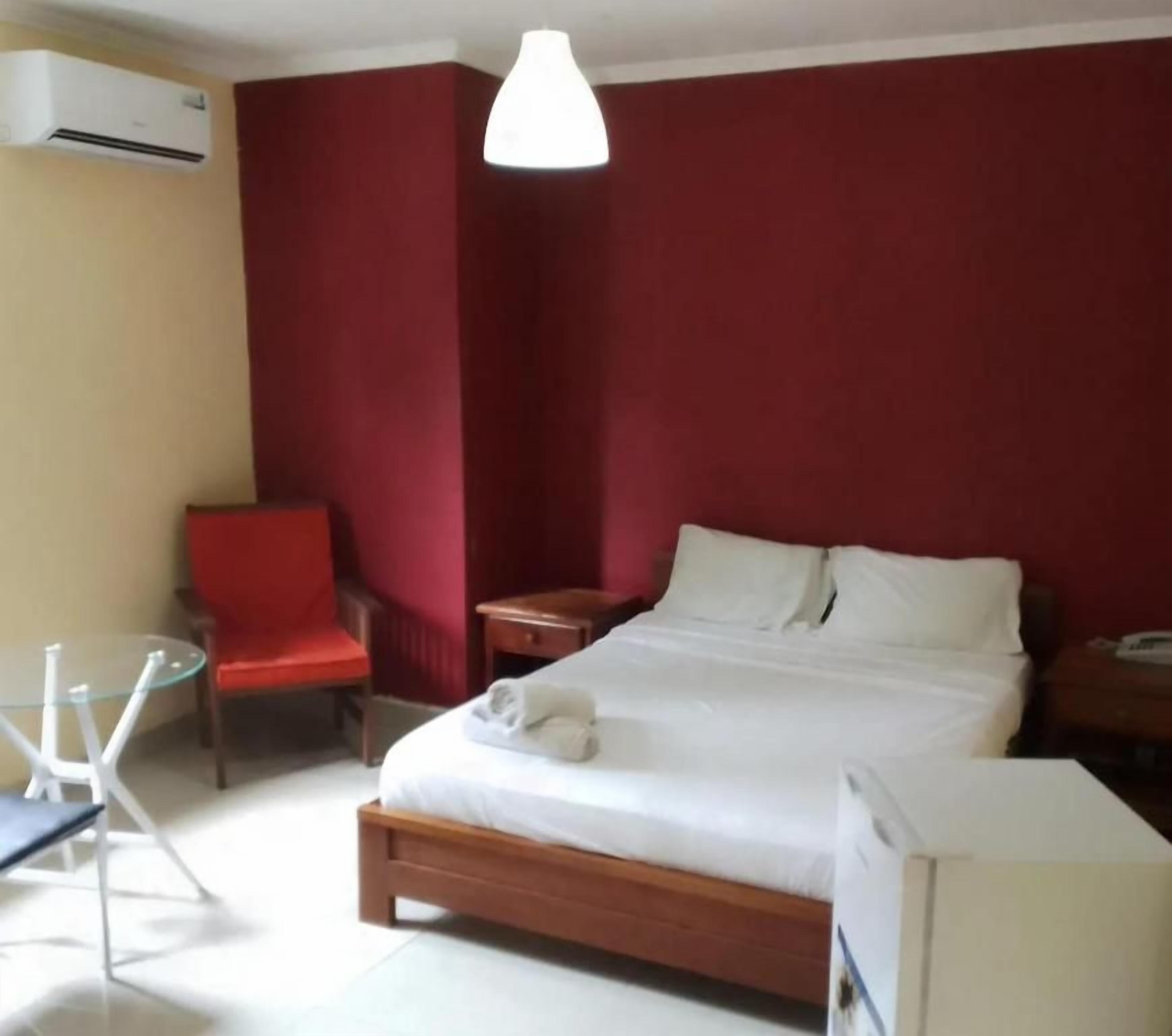 Una habitación de hotel con una cama, una silla roja, una mesita pequeña y una lámpara blanca.