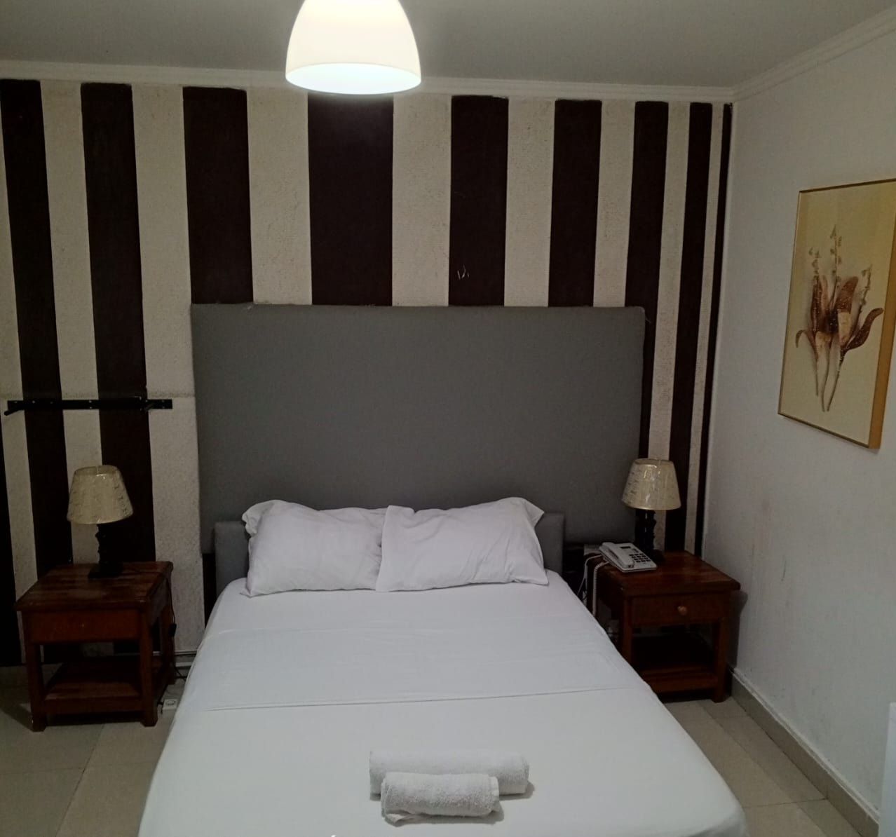 Un cuarto de hotel con una cama grande, dos mesitas de noche, una lámpara sobre cada una y un teléfono en una de ellas, además de un cuadro en la pared.