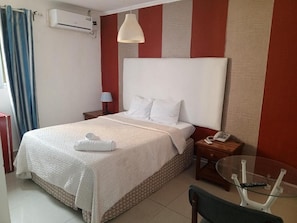 A hotel room with a bed, bedside table, lamp, and a glass table with a remote control. - Angola Stay Connect (Luanda)