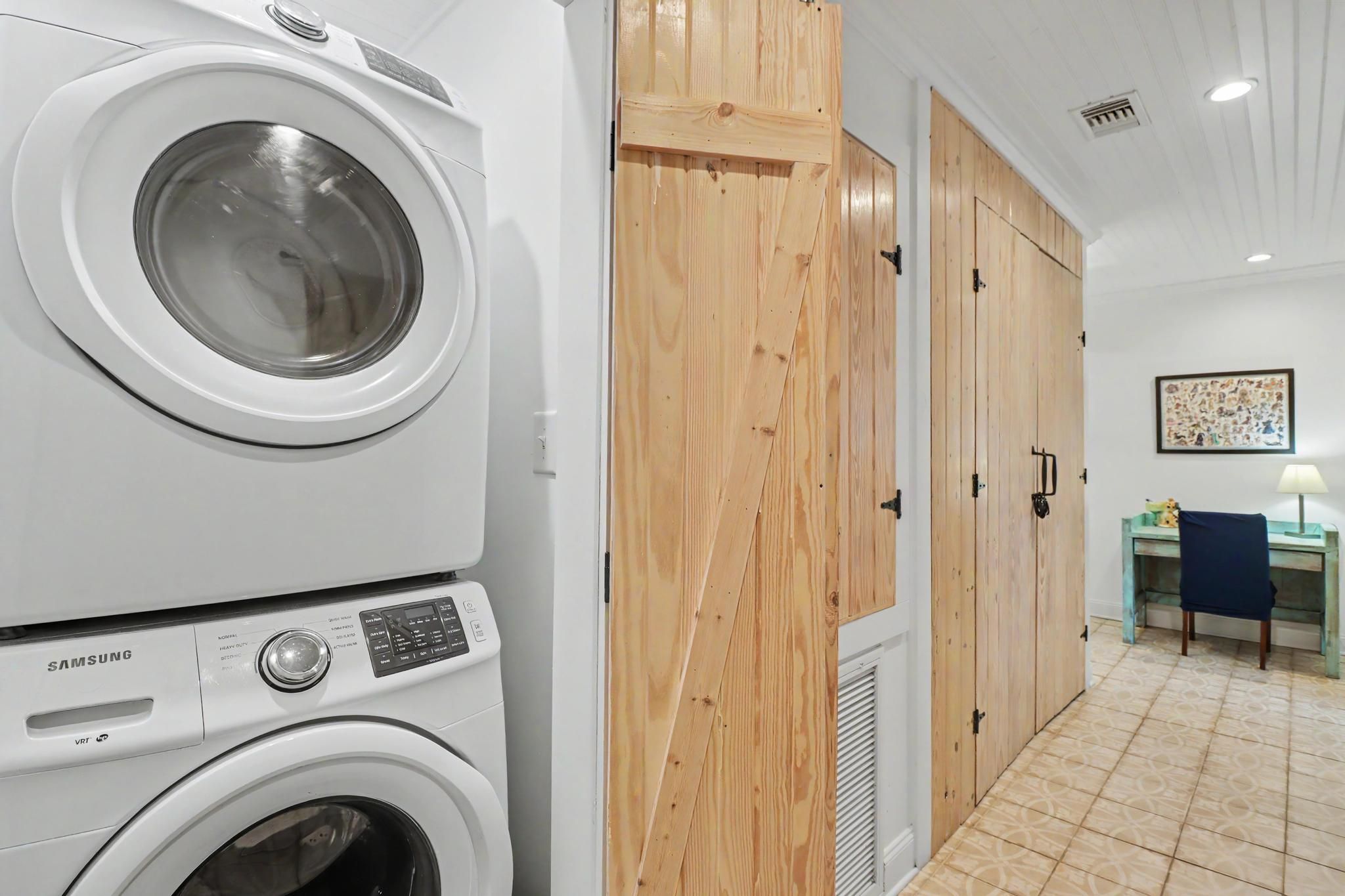A laundry room with stacked washers and dryers, wooden doors, a tiled floor, and a small table with a lamp.