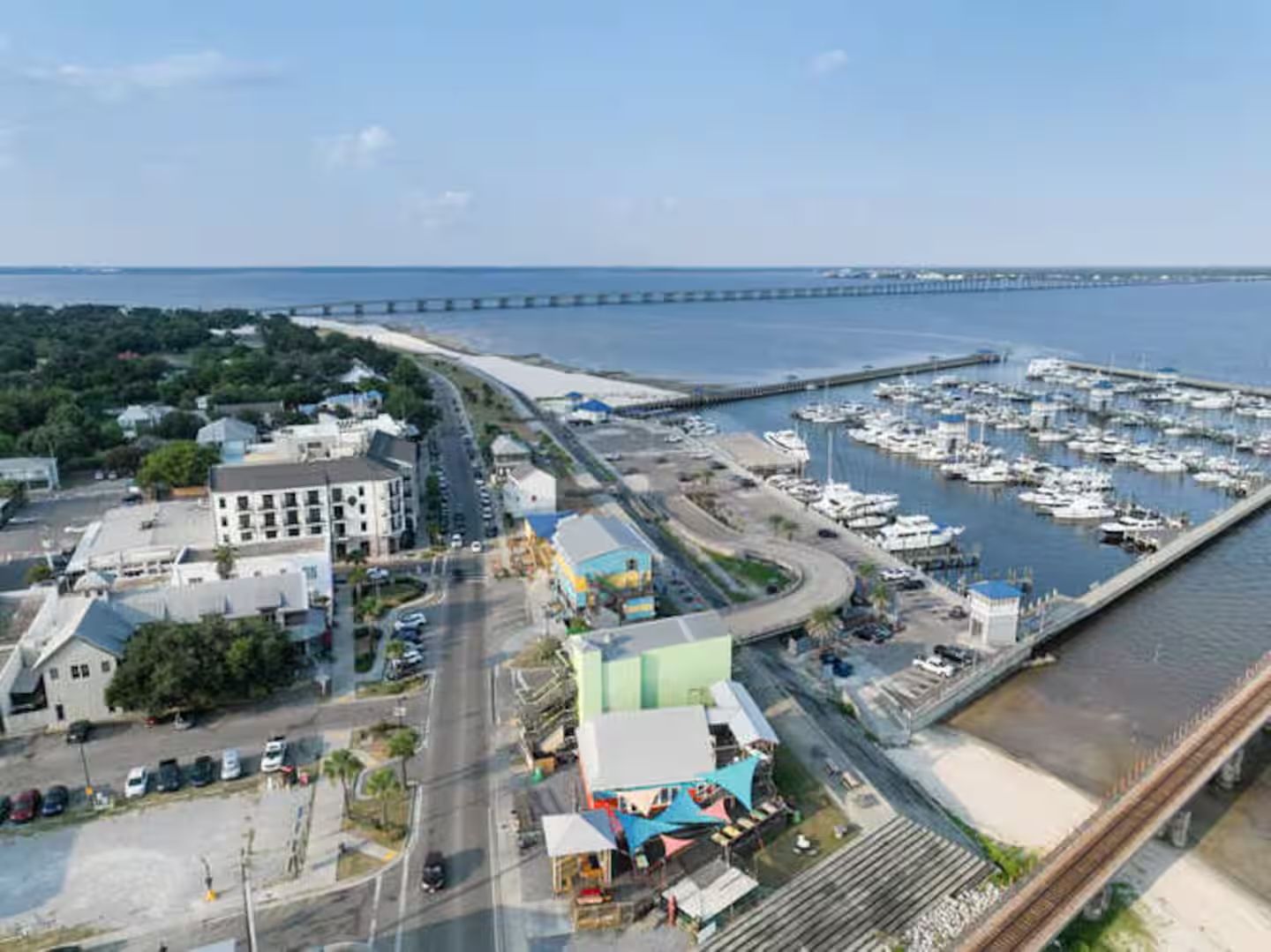 An aerial view of a marina with boats docked, a bridge in the background, and a road leading to a building with a green roof.