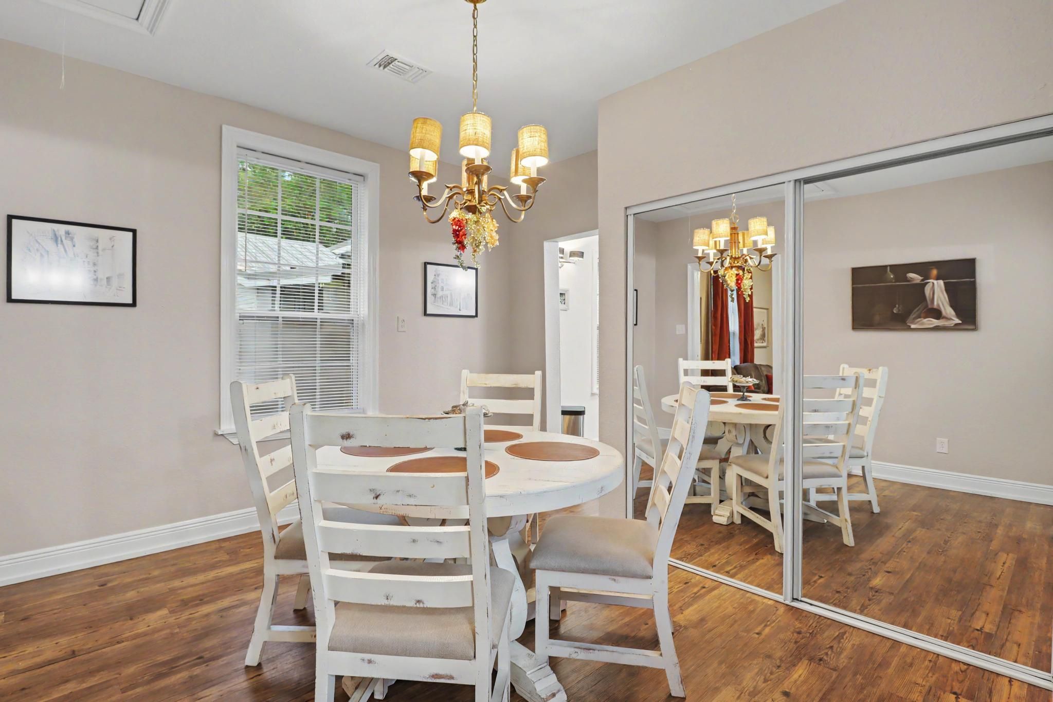 A dining room with a round table, white chairs, a chandelier, and a large mirror reflecting the room.