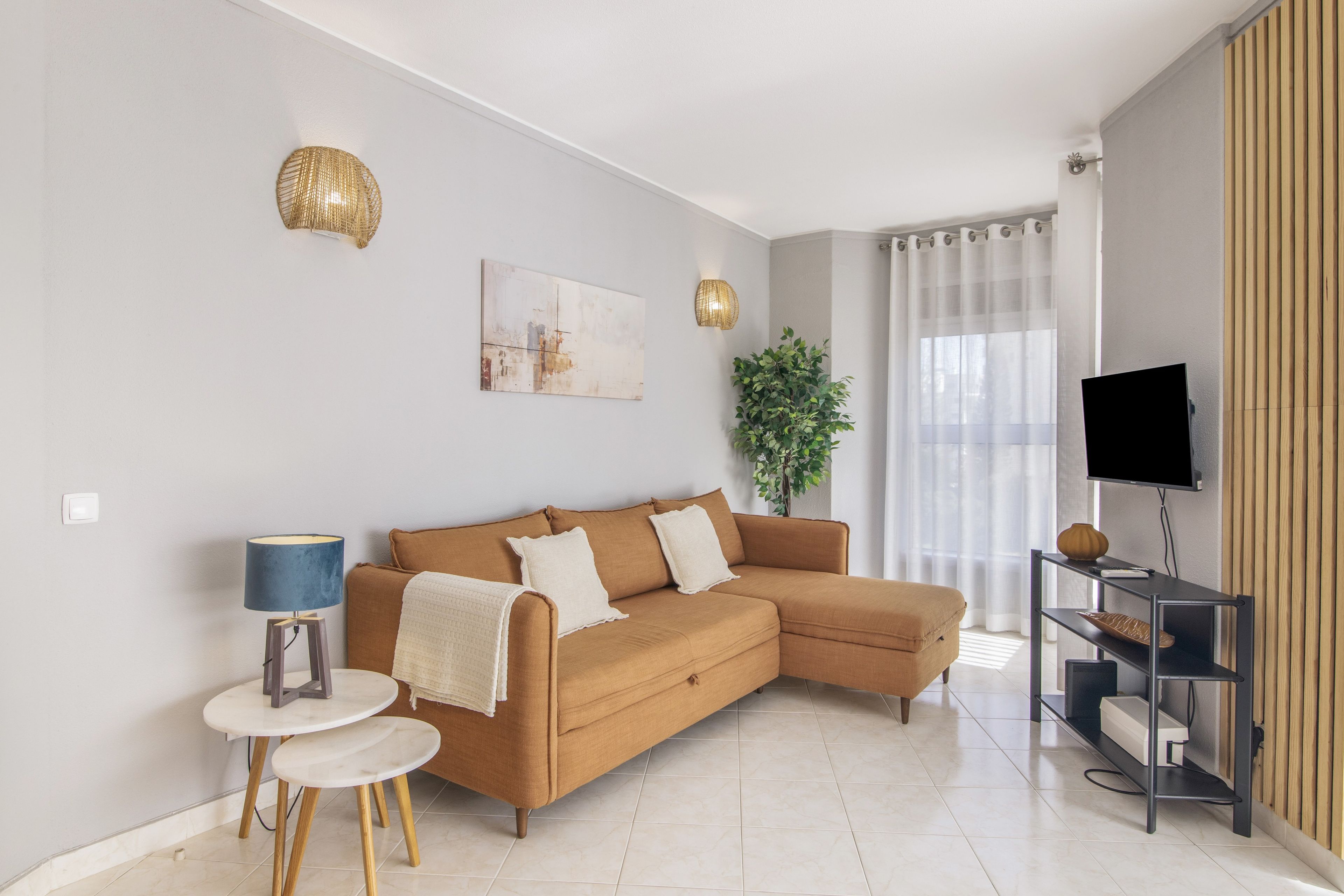Una sala moderna con un sofá seccional, una mesita auxiliar, una lámpara de pie, un televisor sobre un mueble bajo y una planta en maceta.