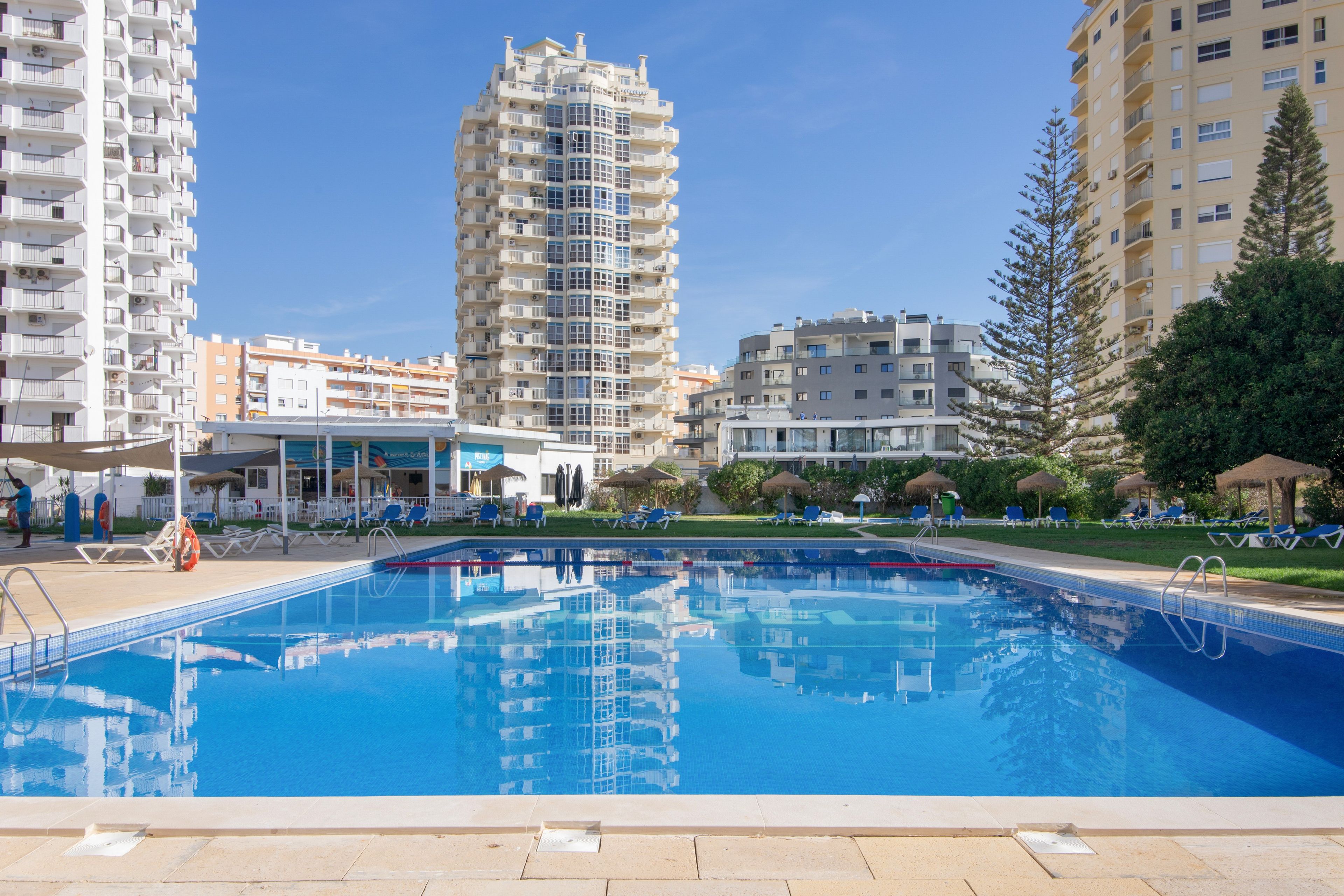 A swimming pool with clear blue water, surrounded by lounge chairs and umbrellas, with high-rise buildings in the background.