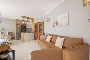 A modern living room with a brown sofa, a dining area with a white table, and a kitchen with wooden cabinets and a built-in oven. - Caprichoso Beachfront Apartment with Shared Pool, Balcony, and Air Conditioning (Armação de Pêra)