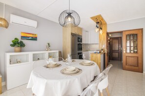 A dining area with a round table set for four, a white cabinet with glass doors, a wooden door, and a kitchen area with modern appliances. - Caprichoso Beachfront Apartment with Shared Pool, Balcony, and Air Conditioning (Armação de Pêra)