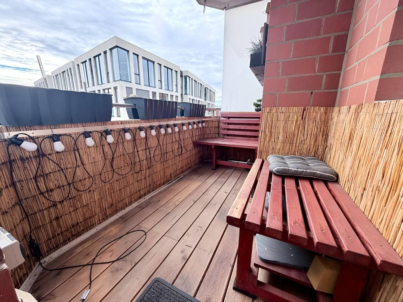 Una terraza de madera con un banco rojo y una mesa, rodeada por una cerca de bambú, con un edificio moderno al fondo.