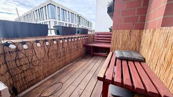 Una terraza de madera con un banco rojo y una mesa, rodeada por una cerca de bambú, con un edificio moderno al fondo.