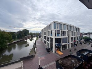 A modern building with a glass facade, a canal in front, and a pedestrian walkway. - City View II Vacation Rental – Terrace & Downtown (Nordhorn)
