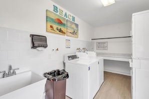 A laundry area with a washing machine, dryer, and a trash bin. There are two paintings on the wall above the washing machine depicting beach scenes. - Sea Club #43 - The Shore Thing (Indian Shores)