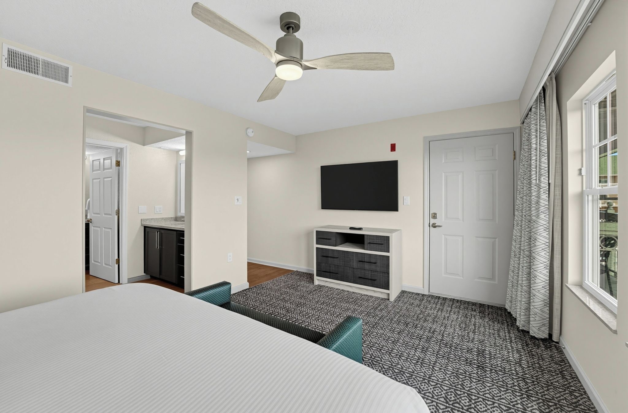 Une chambre à coucher comprenant un lit, un ventilateur de plafond, une télévision, une commode et une fenêtre avec des rideaux.