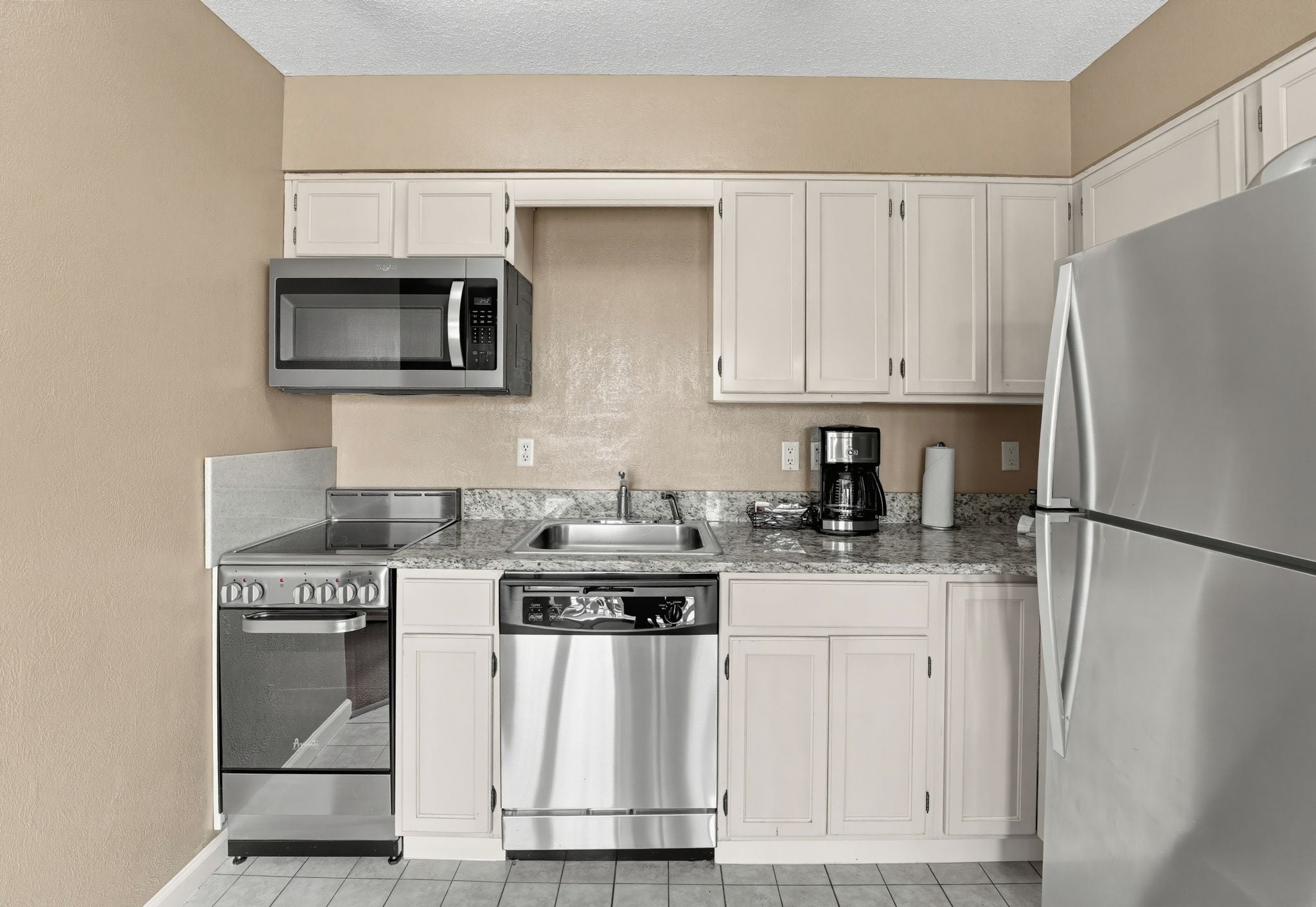 A compact kitchen with white cabinets, a stainless steel refrigerator, a microwave, a dishwasher, and a coffee maker.