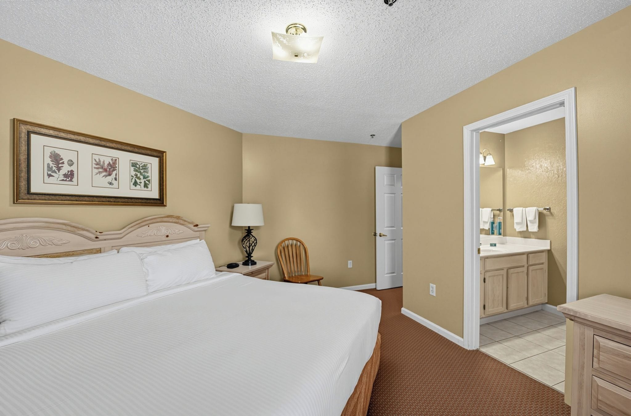 Habitación de hotel con una cama grande, una mesita de noche, una silla, un escritorio, una lámpara, un espejo y un baño visible a través de una puerta abierta.