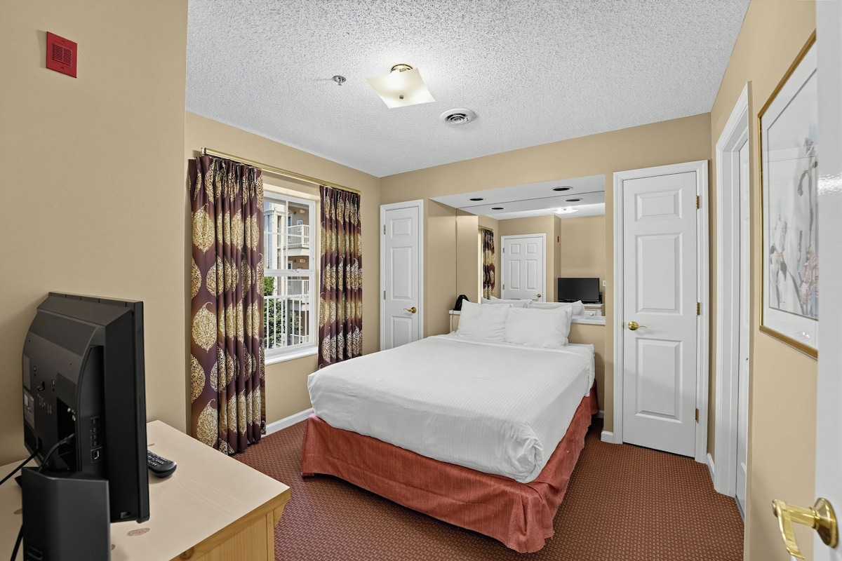 Una habitación de hotel con cama, escritorio con televisor, ventana con cortinas y puerta.