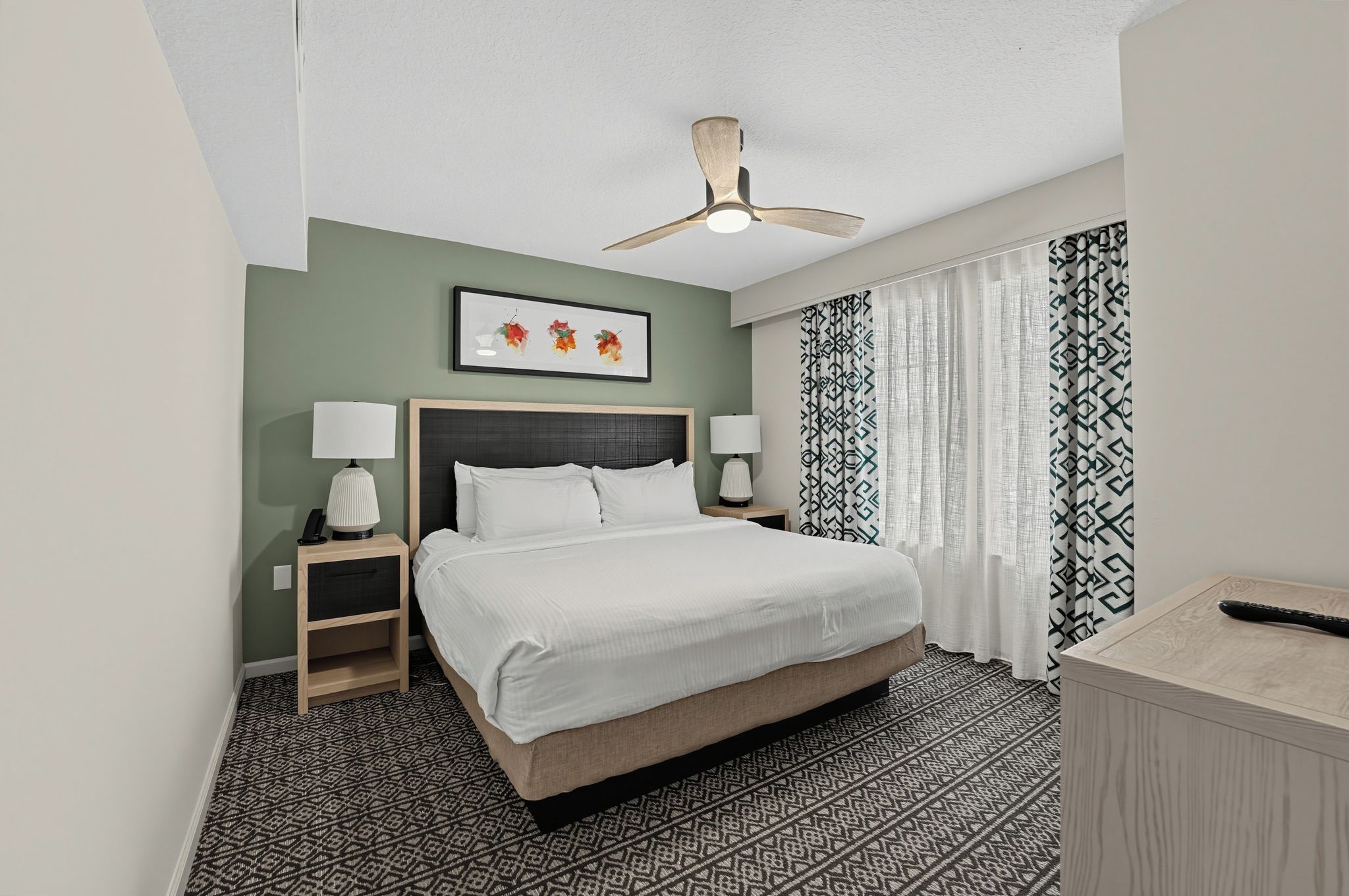 Quarto de hotel com uma cama, duas mesas de cabeceira, um ventilador de teto, uma obra de arte emoldurada, cortinas estampadas e um tapete estampado.