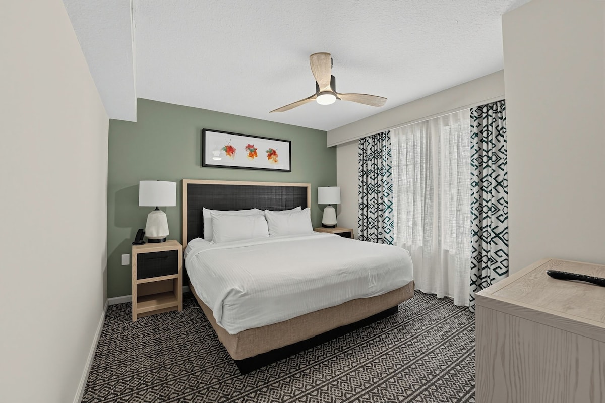 Habitación de hotel con cama, dos mesitas de noche, ventilador de techo, un cuadro enmarcado, cortinas estampadas y alfombra con diseño.