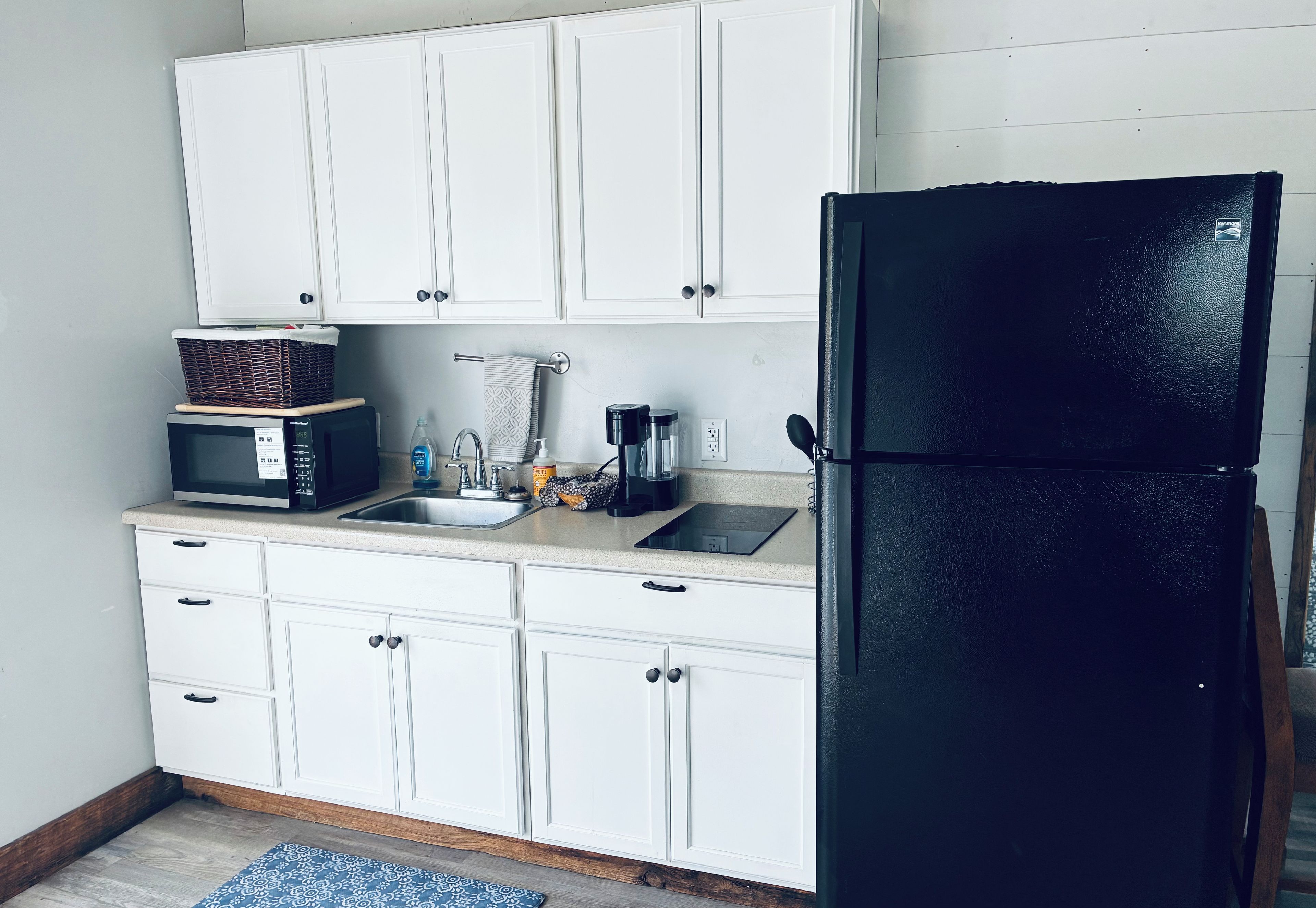 A kitchen with white cabinets, a black refrigerator, a microwave, a sink, and a patterned rug.