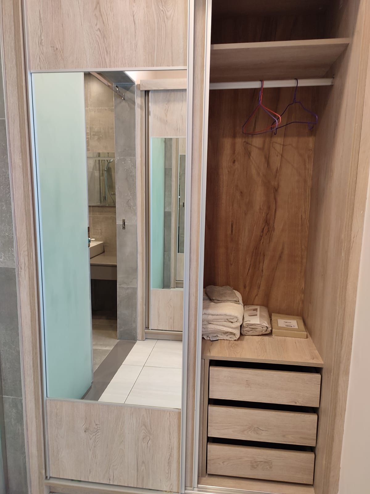 Un dressing accessible sans porte, doté d’une porte de placard avec miroir, d’une étagère en bois où sont pliées des serviettes et d’une tringle pour suspendre les vêtements.