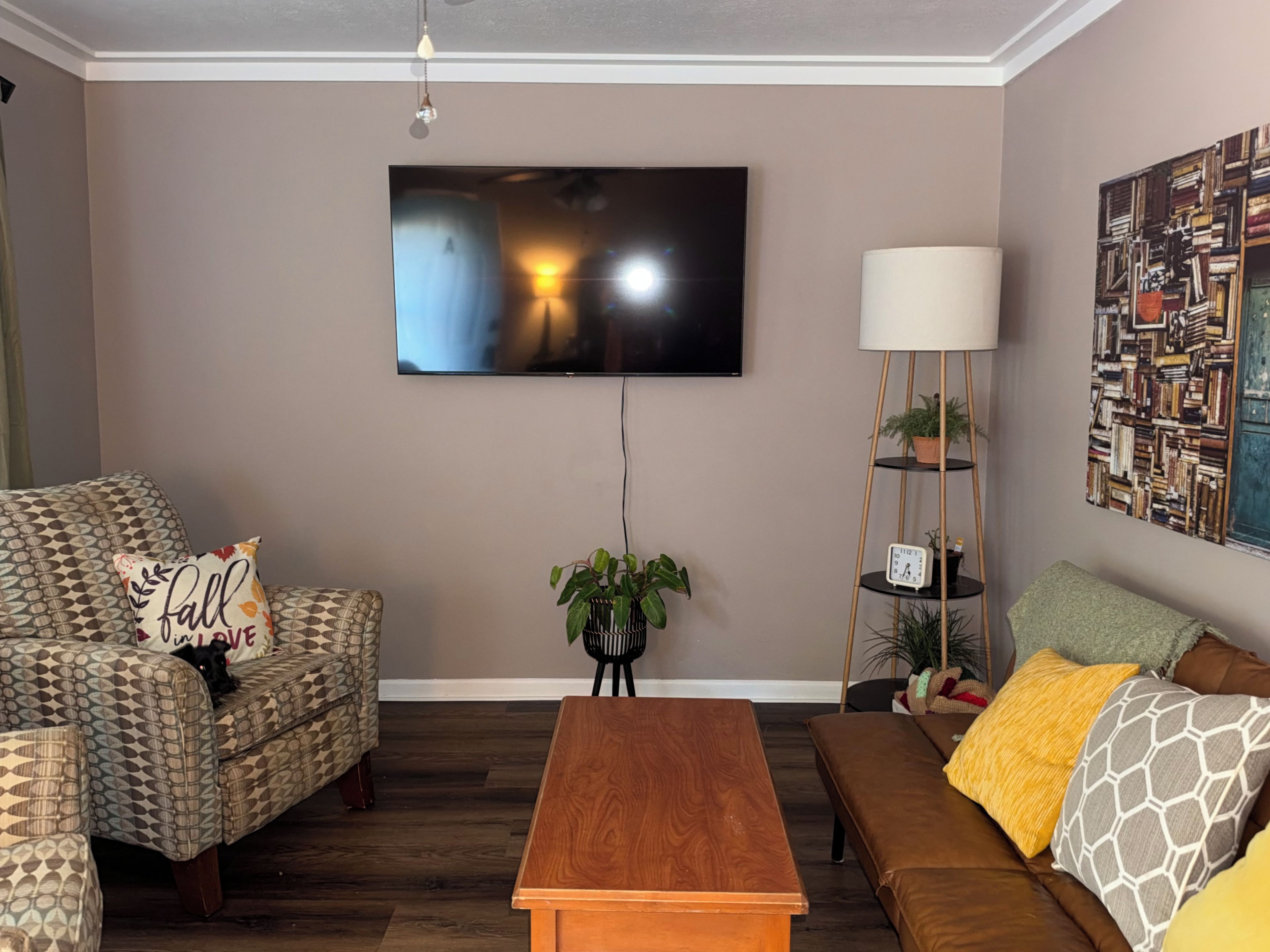 A living room with a flat-screen TV mounted on the wall, a wooden coffee table, patterned sofas, and a bookshelf with a lamp.