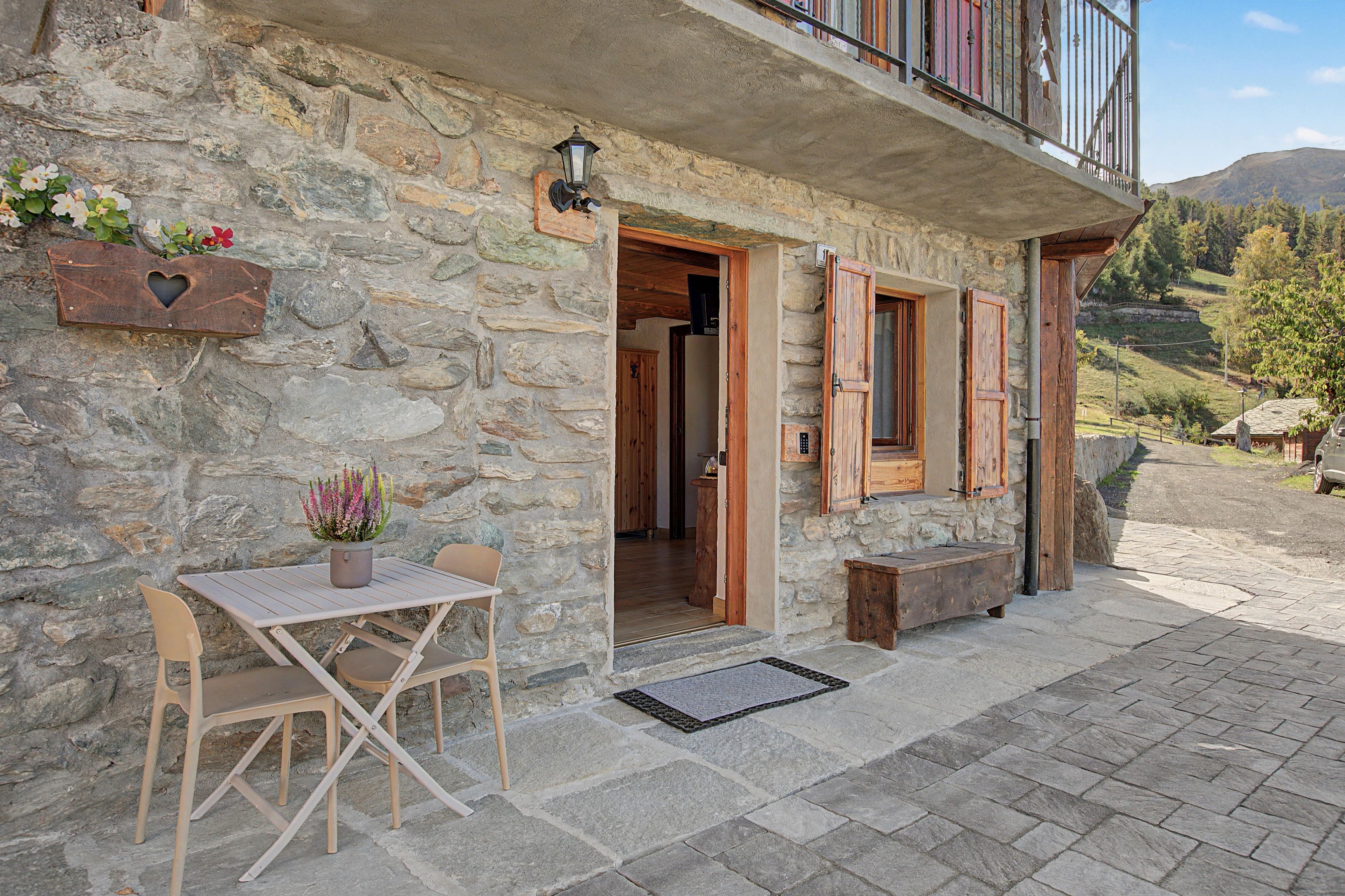 A stone building with a wooden balcony, a table with chairs, and a potted plant.