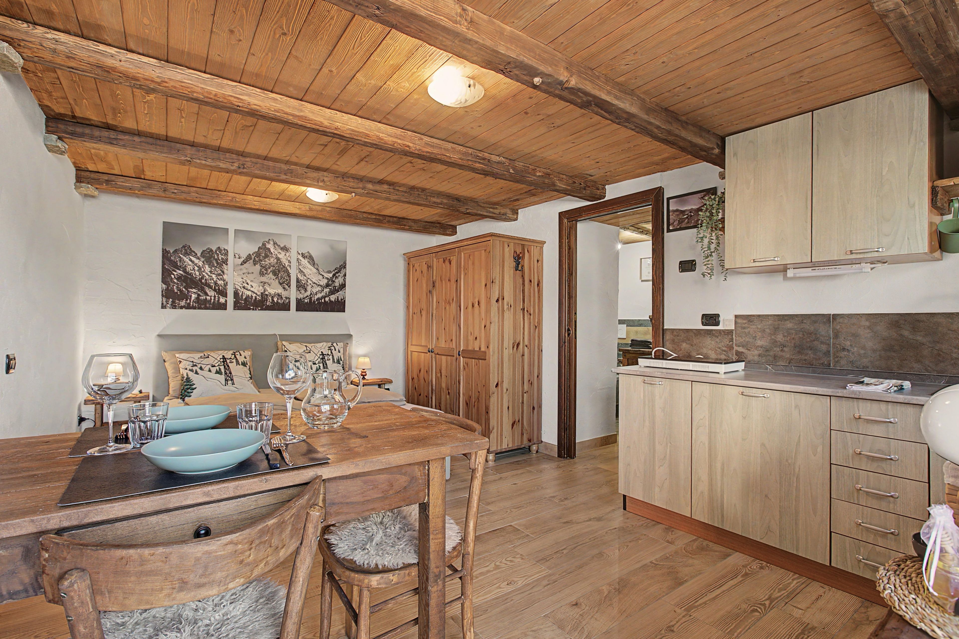 Una mesa de comedor de madera con sillas, una zona de cocina con armarios de madera y un techo con vigas a la vista.