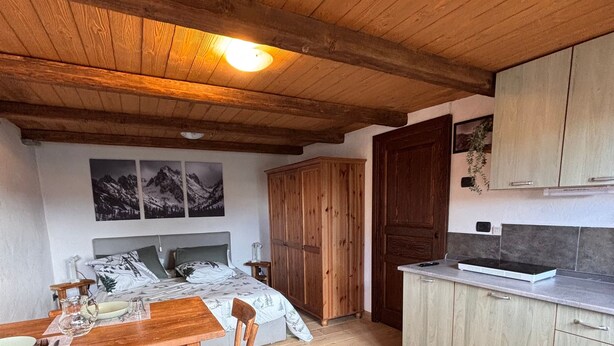 A wooden dining table with chairs, a bed with a white and green patterned bedspread, wooden cabinets, and a tiled floor.