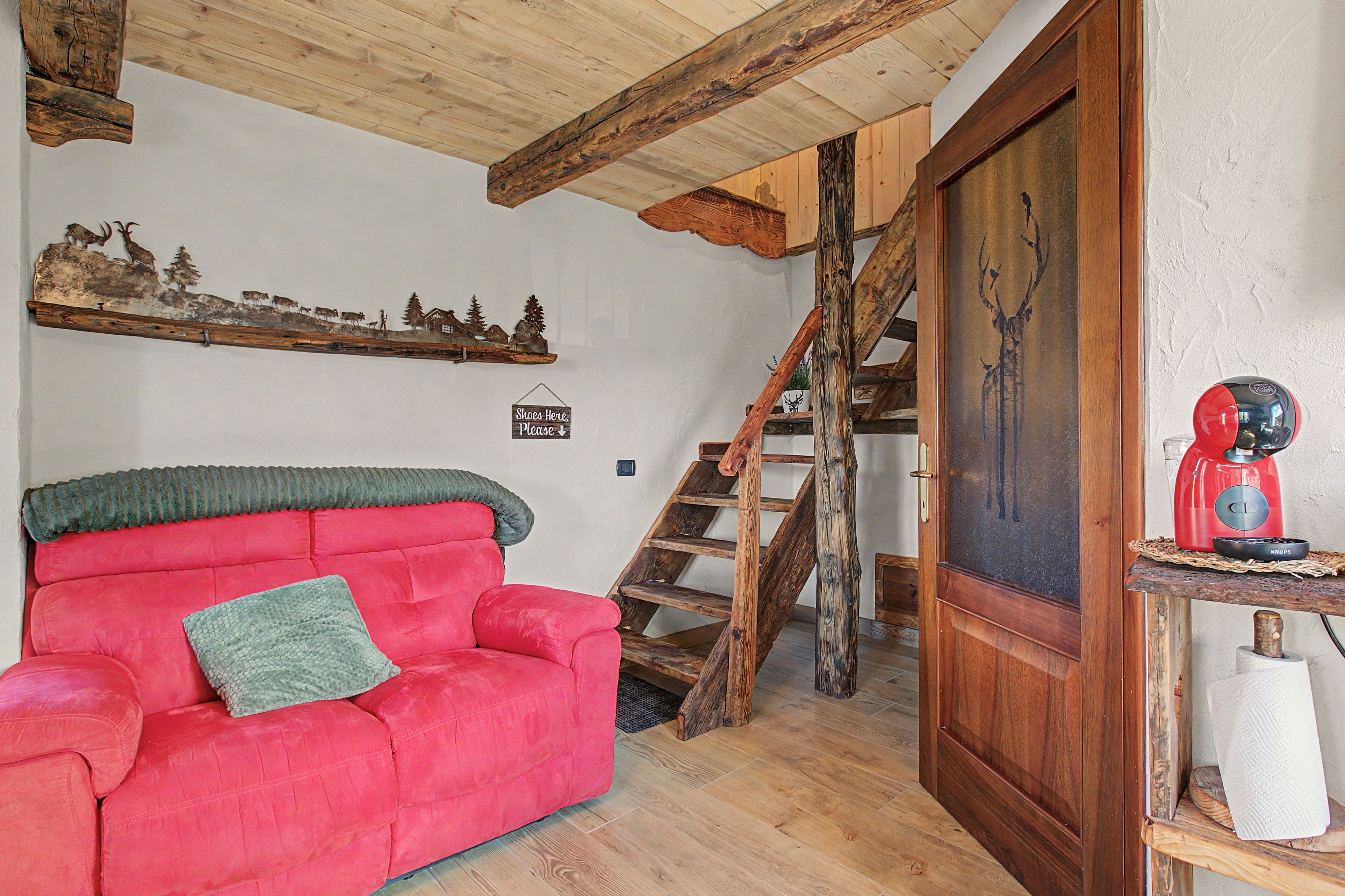 Una habitación acogedora con un sofá rojo, vigas de madera en el techo, una escalera y un electrodoméstico rojo sobre una estantería de madera.