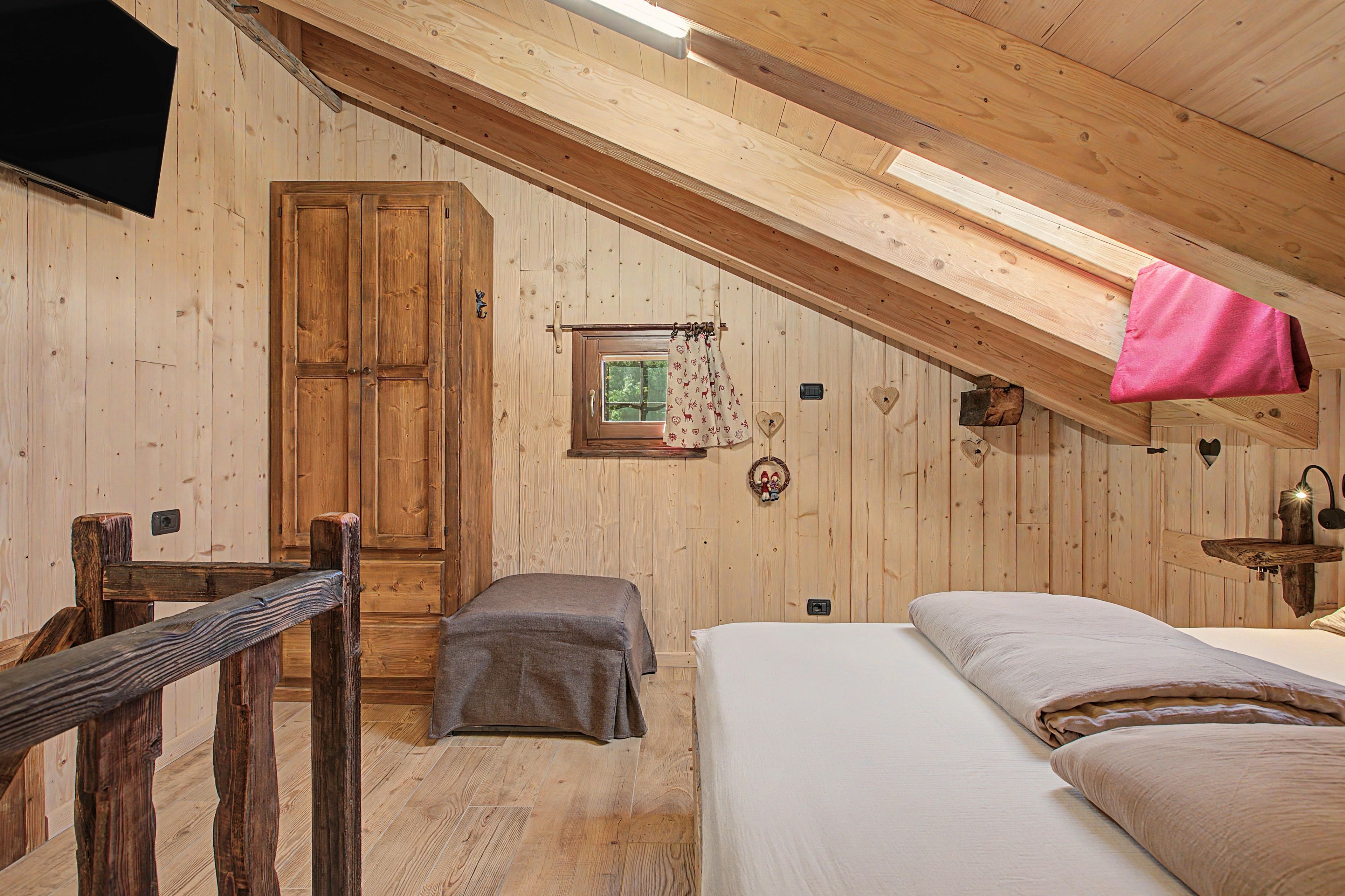 A wooden room with a bed, a bench, a wardrobe, and a window with a curtain.