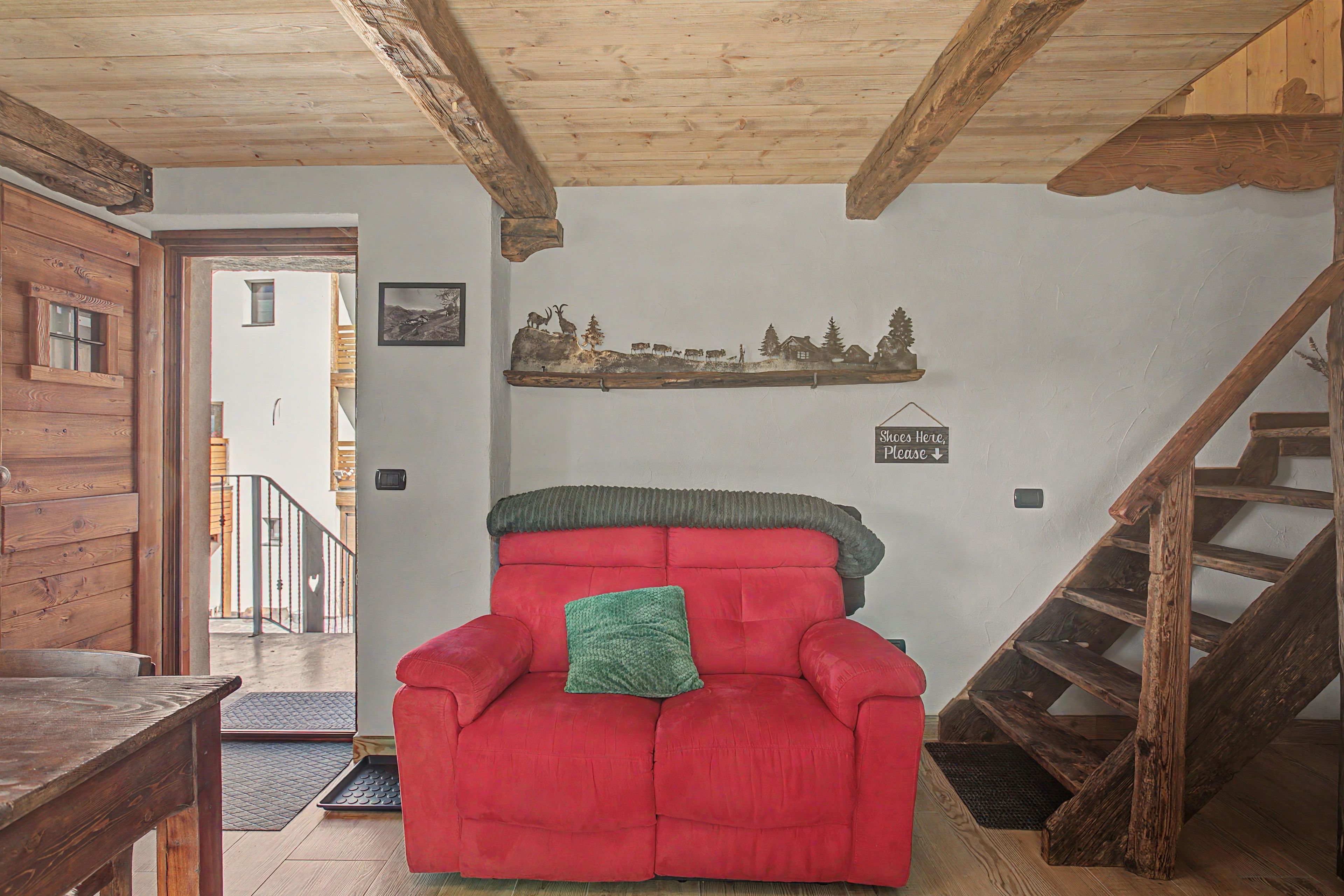 A red armchair with a green pillow in a room with wooden beams, a staircase, and a wall-mounted shelf with a landscape picture.